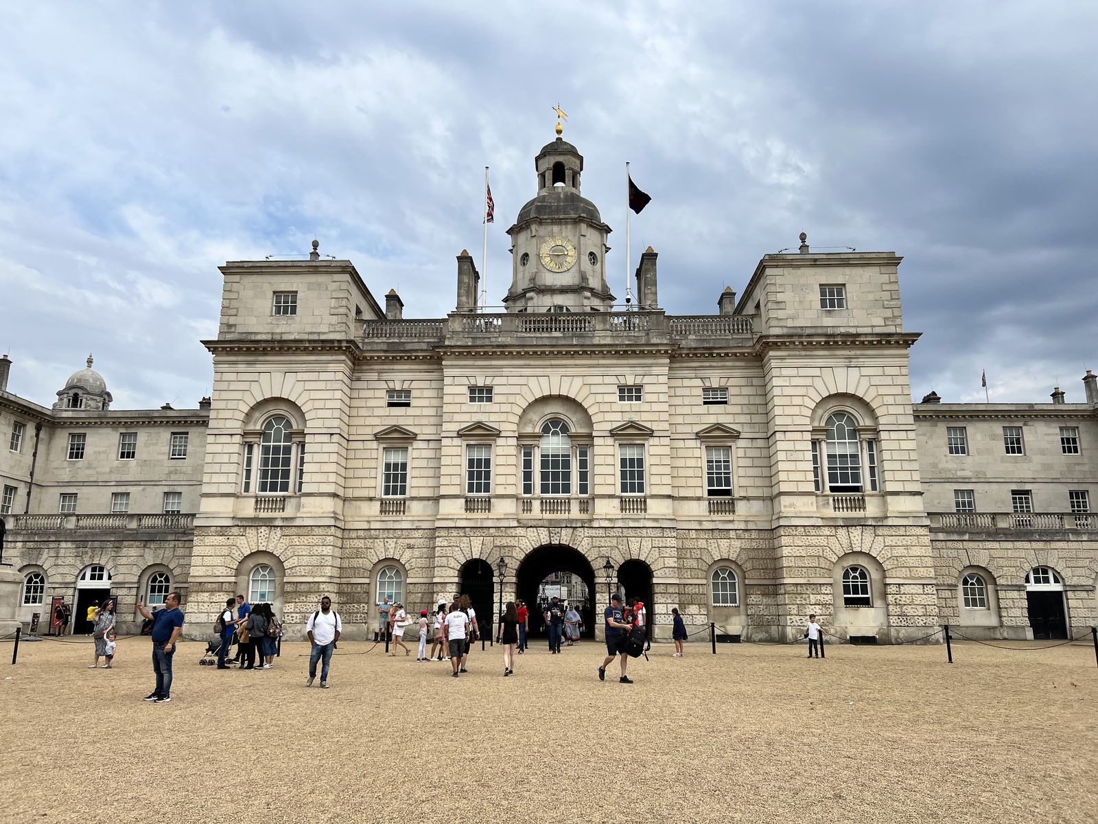 Historic stone building under cloudy sky, people walking and gathering outside. Clock tower centered, flag atop.