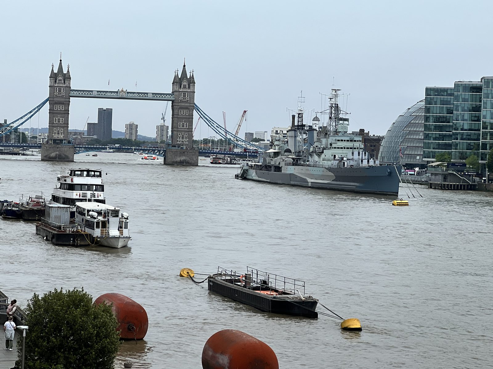 Tower Bridge over the Thames, with boats docked nearby. A warship is moored, and modern buildings are visible.