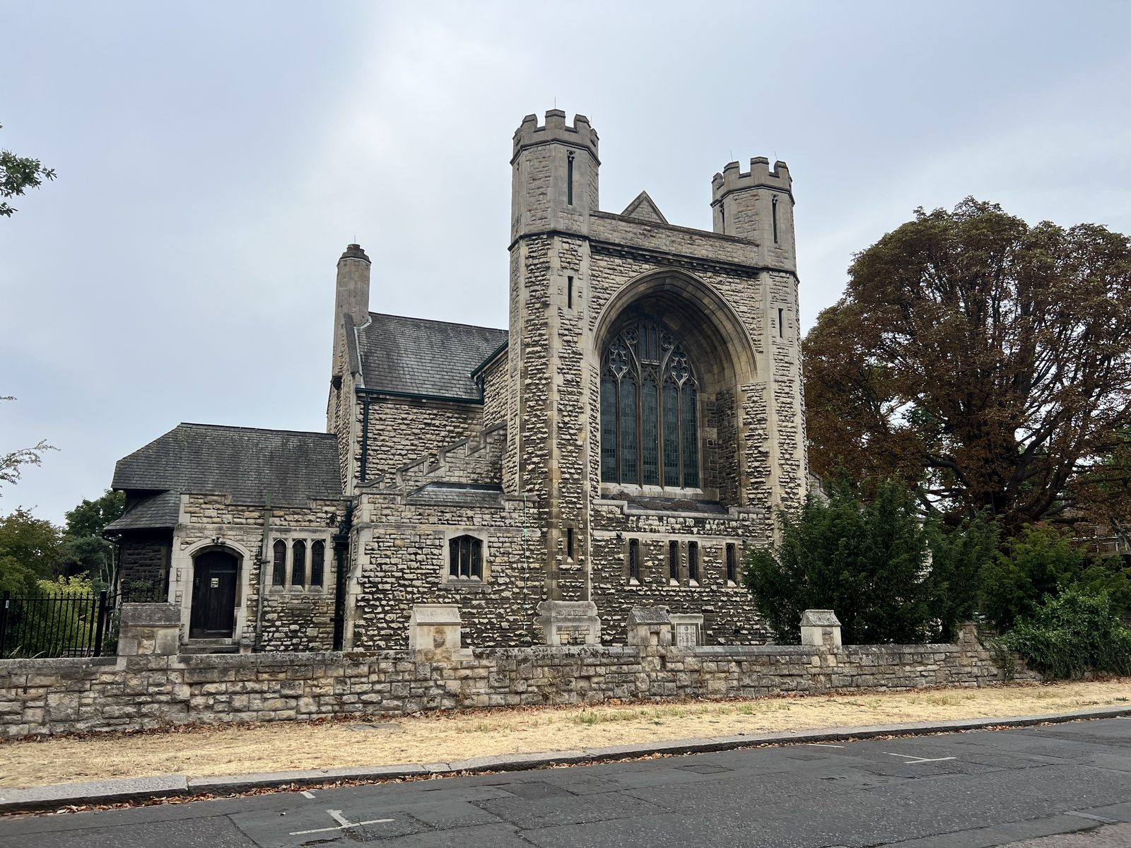 Stone church with tall arched window, flanked by trees and a stone wall.